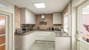 A compact kitchen with white cabinets, a brick accent wall, a stainless steel range hood, and a window with a view of the beach. - Host & Stay | The Braes (Marske-by-the-Sea)