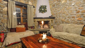 A cozy room with a stone wall, a fireplace, a bed with a red blanket, a fluffy sofa, and a wooden table with candles.