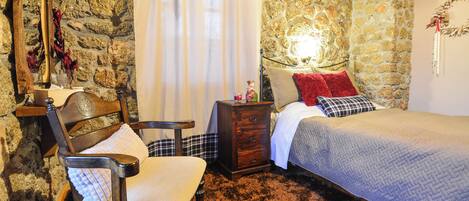 A bedroom with a stone wall, a wooden chair, a bed with a red pillow, and a nightstand.