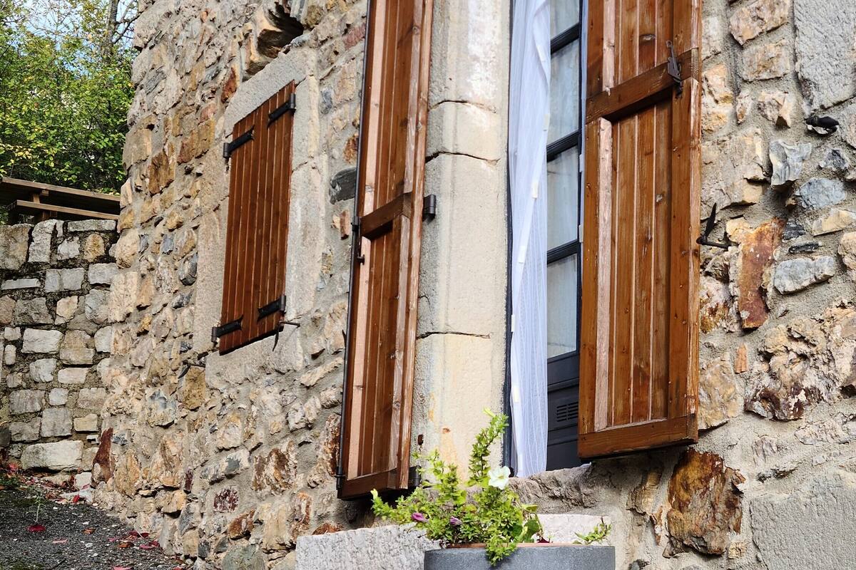 A stone building with wooden shutters, a small window with a curtain, and a potted plant in front.
