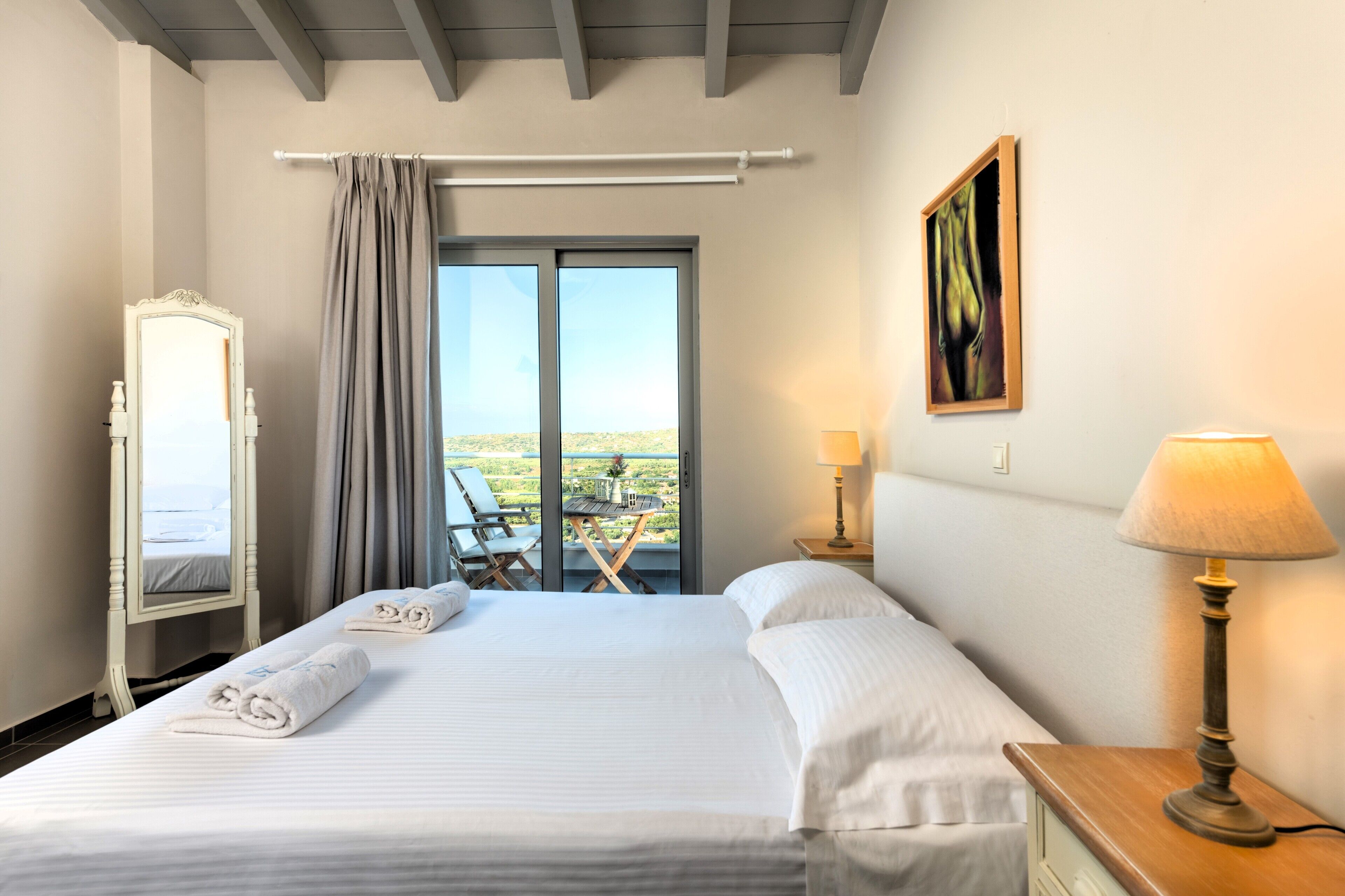 A hotel room with a large bed, a window with a view, a chair, and a table outside.