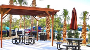 A covered outdoor seating area with tables and chairs, surrounded by palm trees and parked cars. - Charming 1BR/1B~sleeps 6~Gym, Pool, & Boat Parking (Orange Beach)