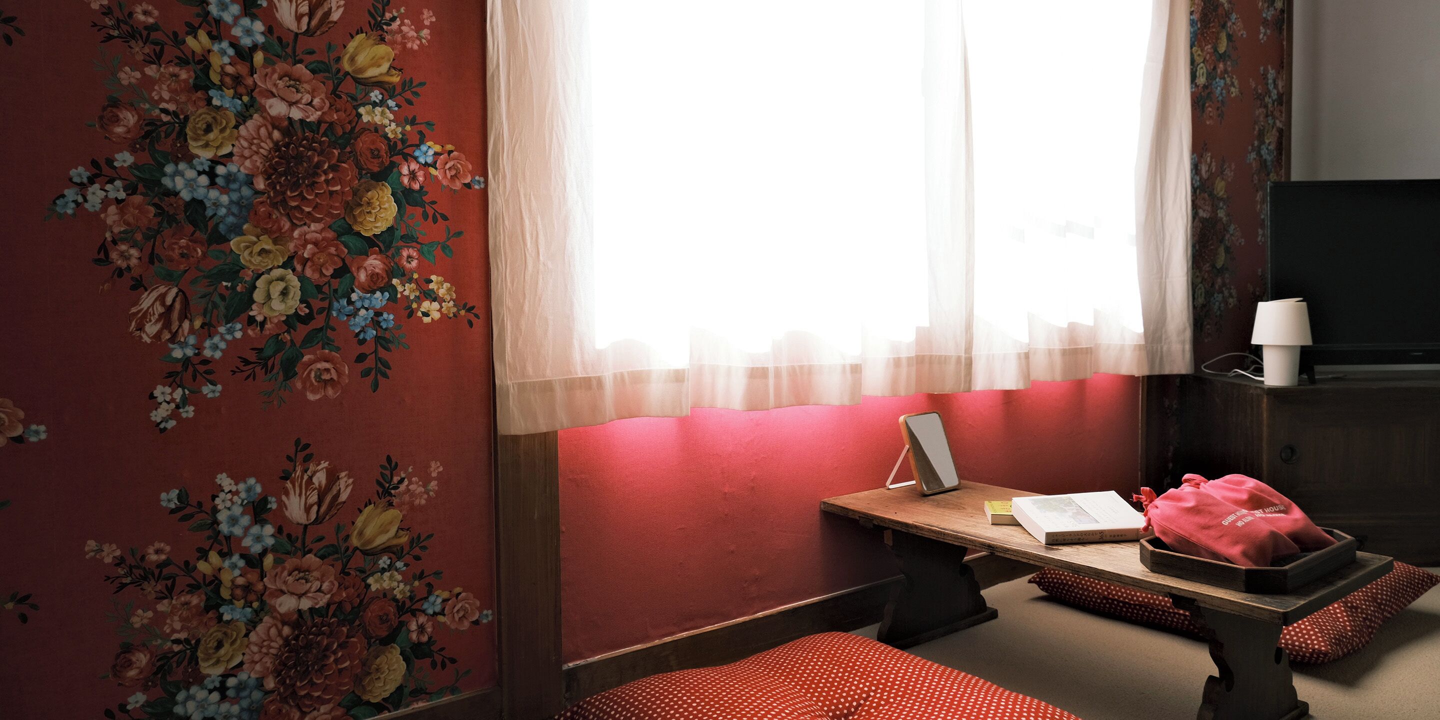 Una habitación con papel tapiz floral, un escritorio de madera con una lámpara, una cama con manta de lunares y un televisor sobre un mueble.