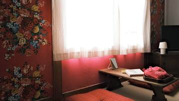 Una habitación con papel tapiz floral, un escritorio de madera con una lámpara, una cama con manta de lunares y un televisor sobre un mueble.