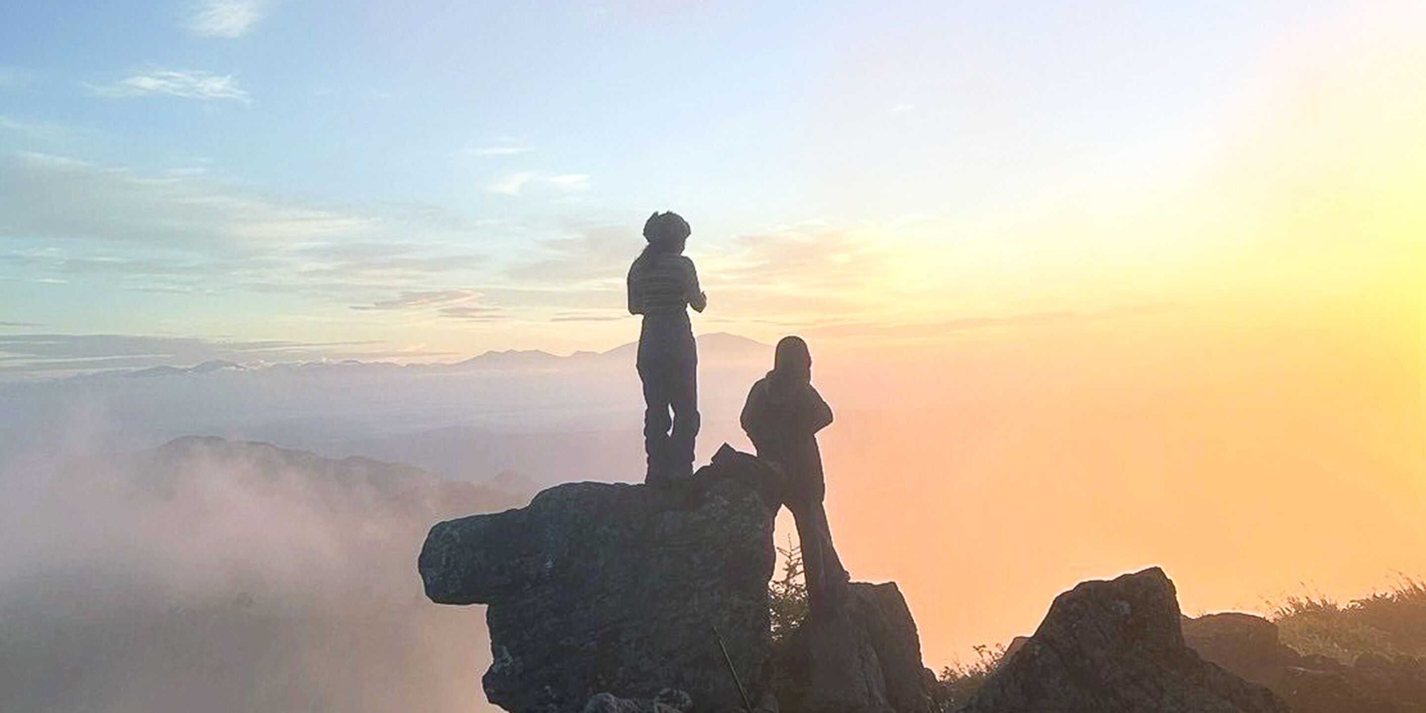 Deux personnes se tiennent debout sur un promontoire rocheux, d’où l’on aperçoit des montagnes lointaines, avec un lever ou un coucher de soleil en arrière-plan.
