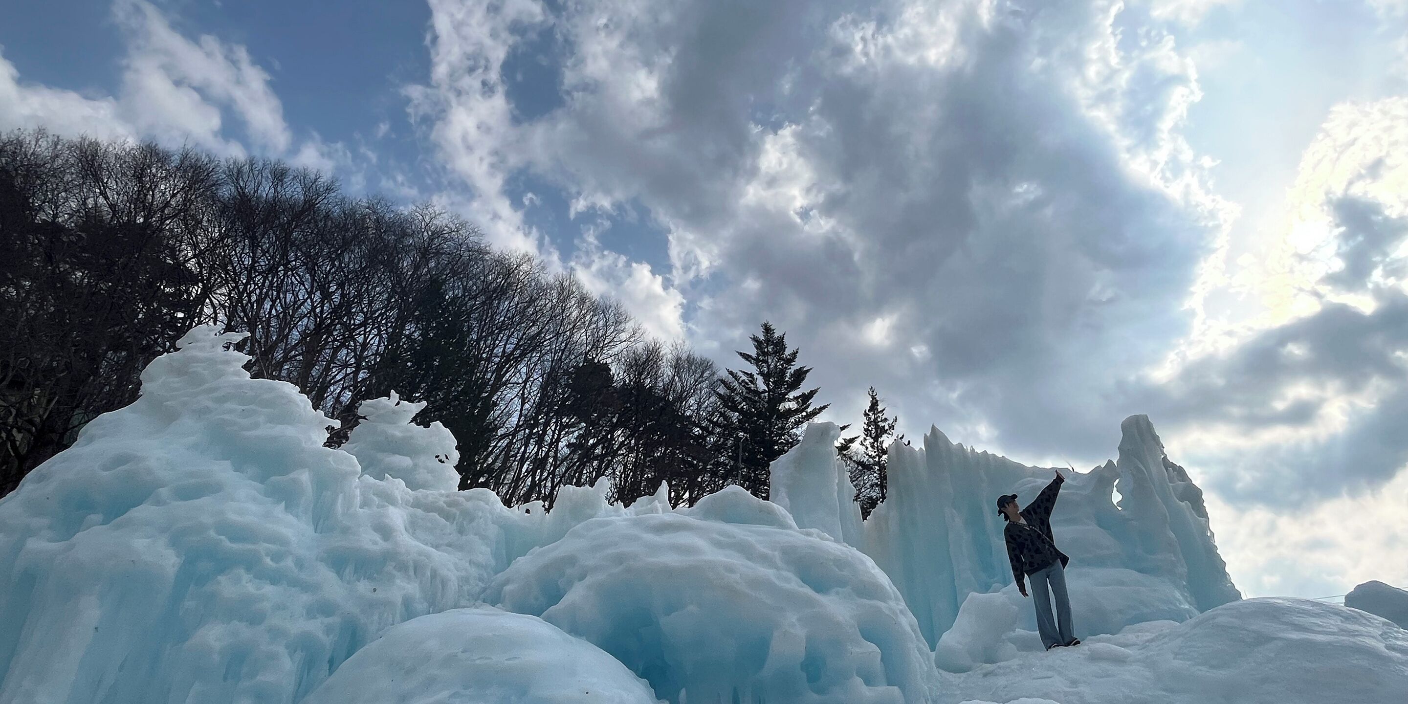 Eine Person steht auf einer schneeüberzogenen Eisskulptur, im Hintergrund sind Bäume zu sehen.