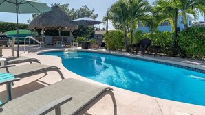 En swimmingpool med liggestole og parasoller, omgivet af palmer og en stråtækt bygning.