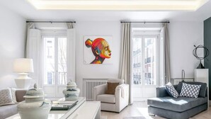 Una sala de estar moderna con un sofá, una mesa de centro y una silla. Hay una ventana con cortinas, un radiador y un cuadro en la pared.