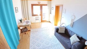 A living room with a sofa, a dining table, and a chair. There is a window with curtains, a ceiling with wooden beams, and a wall with a picture frame. - Die Labsalerie am Wörthersee - Apartment with 1 bedroom (Pörtschach am Wörthersee)