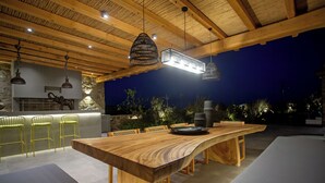 Een terras met een grote houten tafel en stoelen, een bar met krukken en hangende hanglampen.