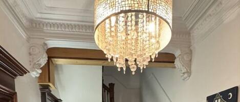 A hallway with a wooden staircase, a chandelier, a glass table with books and a bottle, and a painting on the wall.