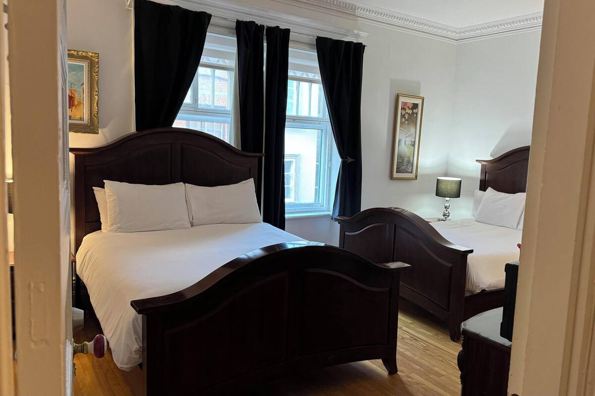 A hotel room with two dark wooden beds, white bedding, a nightstand with a lamp, and a window with black curtains.