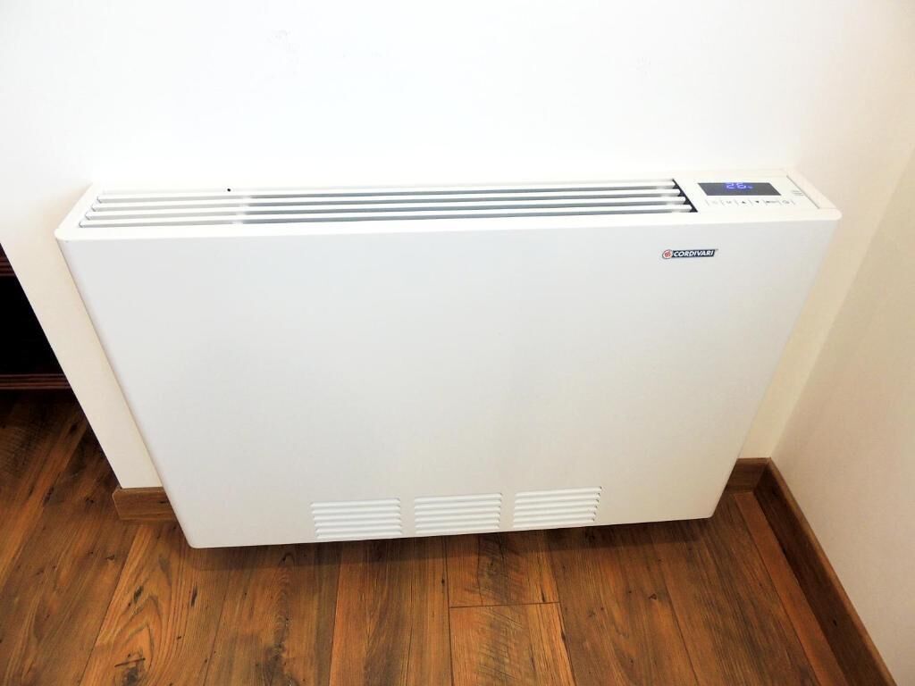 A white portable heater with a digital display and control panel, placed on a wooden floor against a wall.