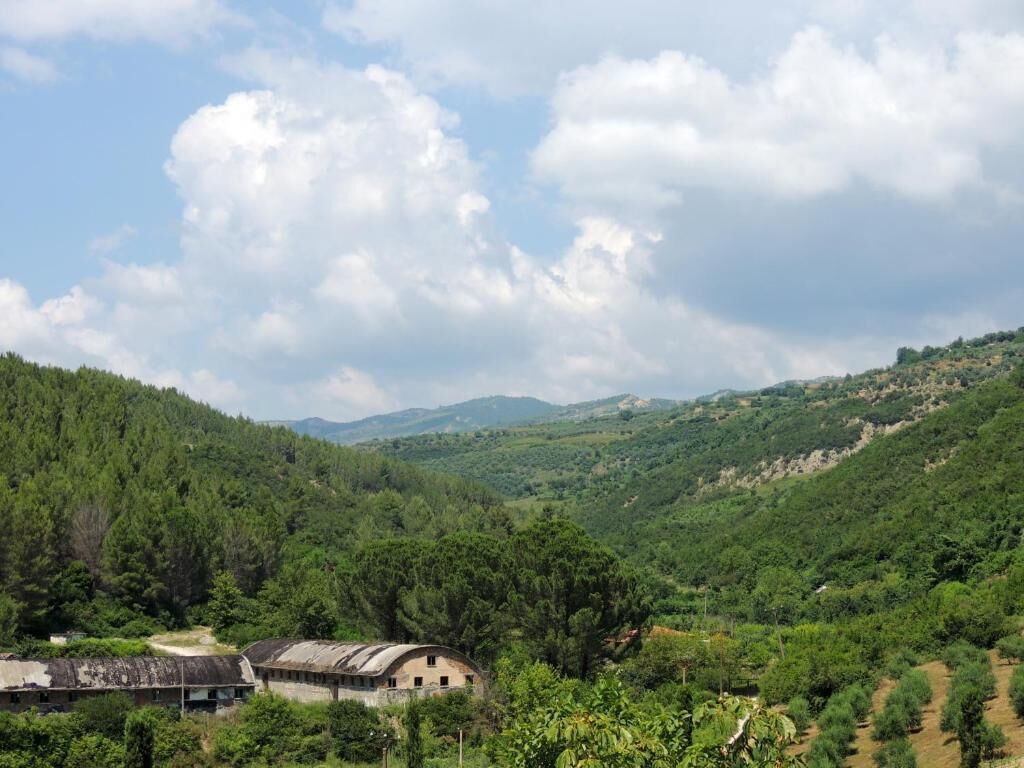 A rural landscape with a large, curved building surrounded by dense forest and rolling hills.