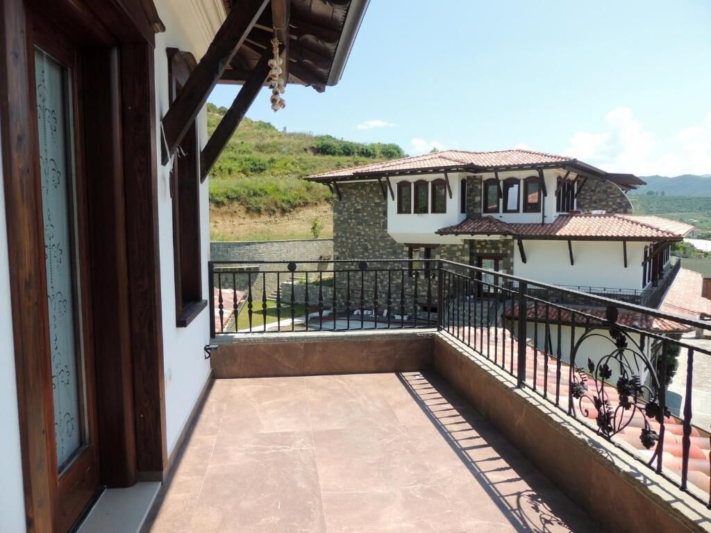 Un balcon avec un sol en pierre, une vue sur une maison avec un toit en tuiles et, au loin, un paysage vallonné.