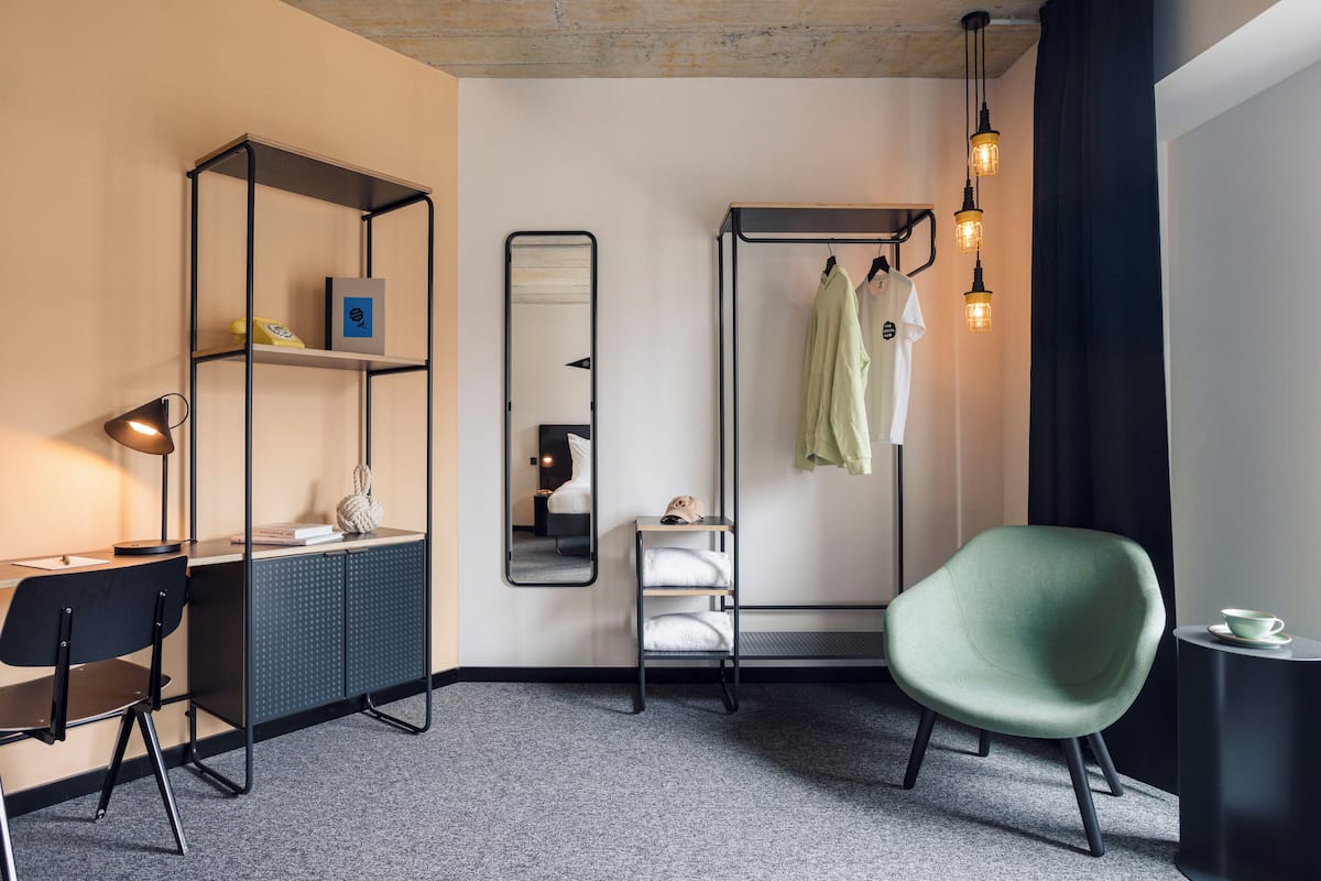 A modern hotel room with a desk, chair, wardrobe, and a green armchair.