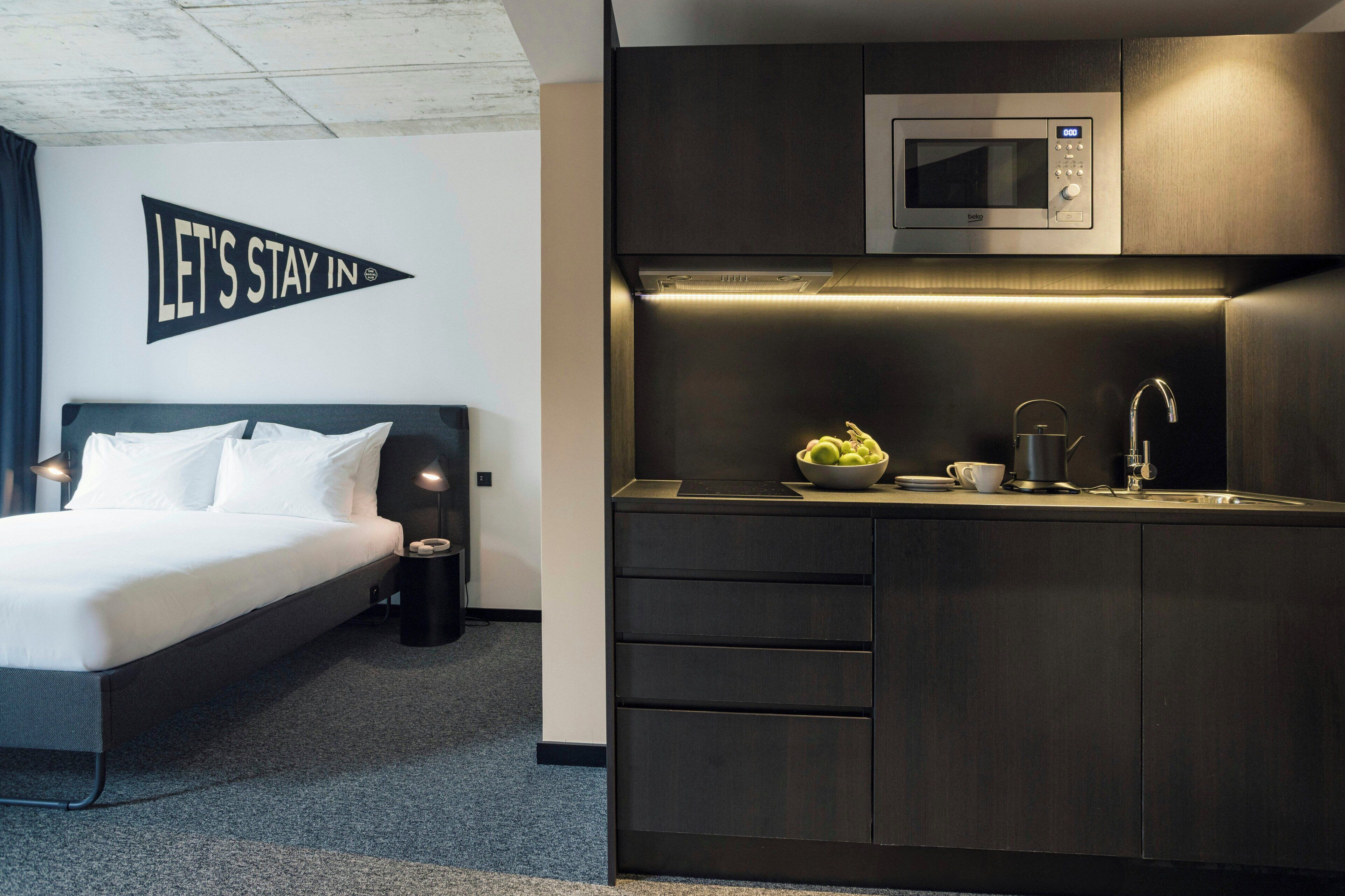 A modern hotel room with a bed, a kitchenette, and a 'LET'S STAY IN' sign.