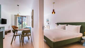 A modern hotel room with a bed, a desk with a chair, a dining table, and a view of the outside.
