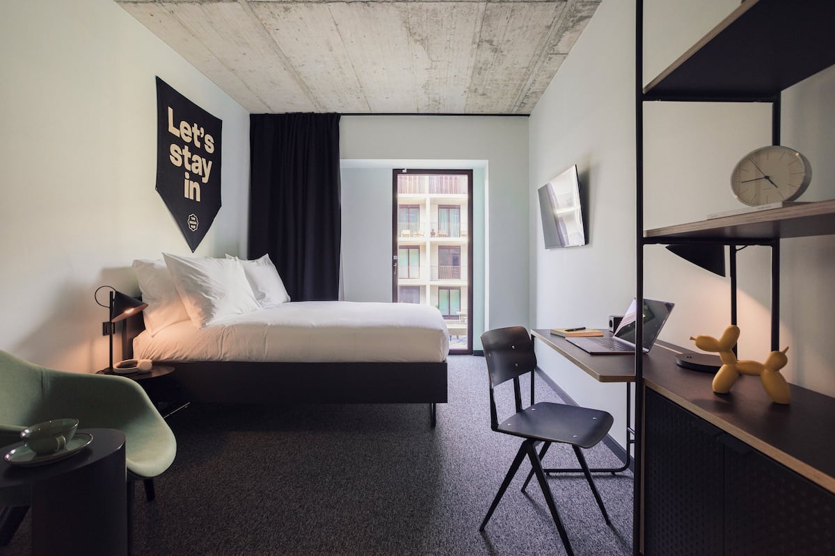 A hotel room with a bed, a desk with a laptop, a chair, a small table with a bowl, and a wall clock.