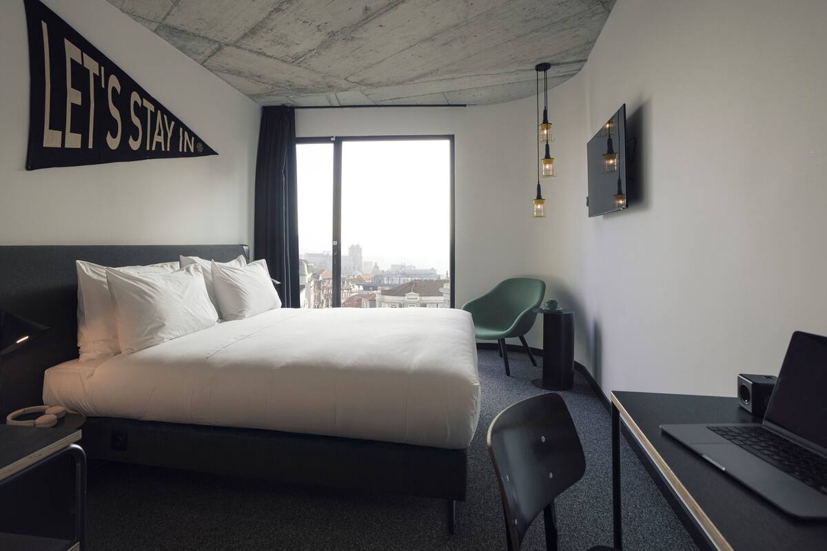 A modern hotel room with a large bed, a desk with a laptop, a green chair, and a city view through the window.