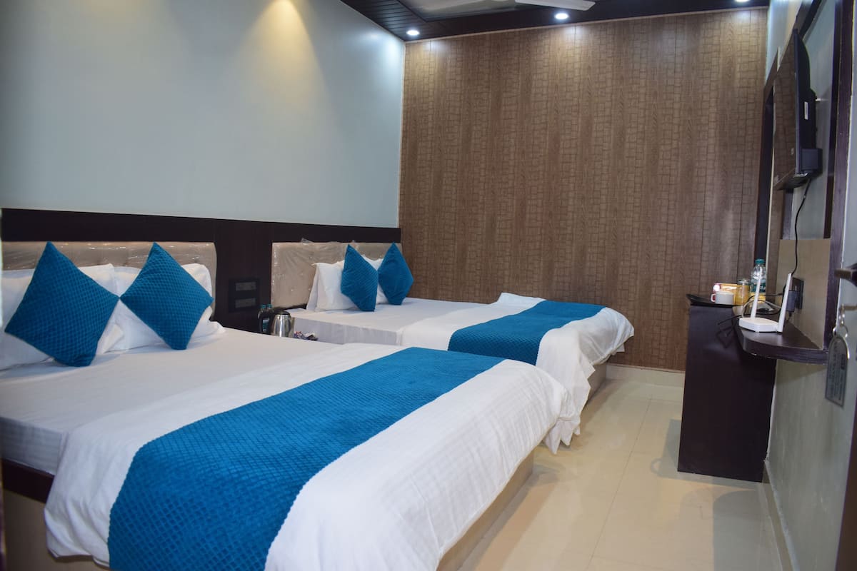 Habitación de hotel con dos camas, cada una con ropa de cama azul y blanca, un panel de pared de madera, un escritorio pequeño con un teléfono y una taza, y un televisor de pantalla plana montado en la pared.