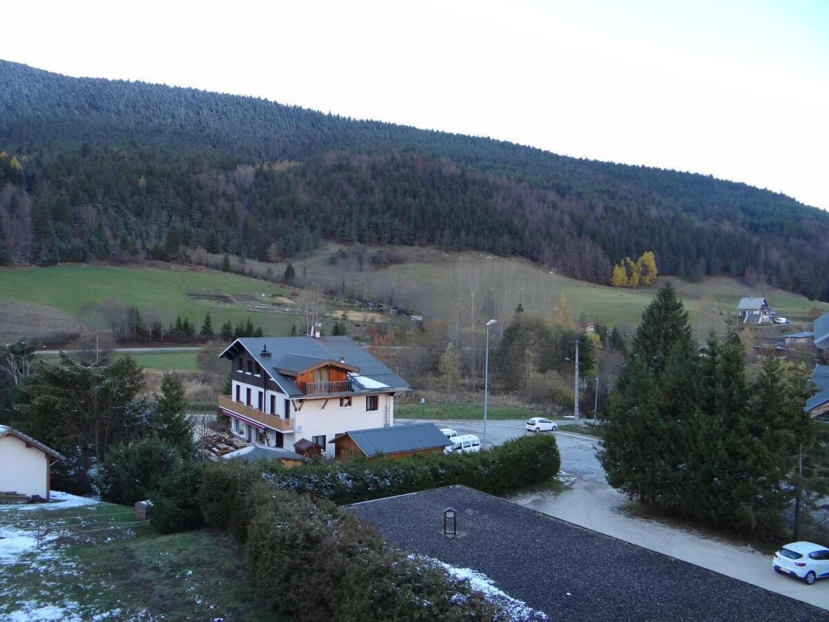 A residential area with a two-story house featuring a balcony, surrounded by trees and a hilly landscape in the background.