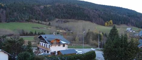 A residential area with a two-story house featuring a balcony, surrounded by trees and a hilly landscape in the background.