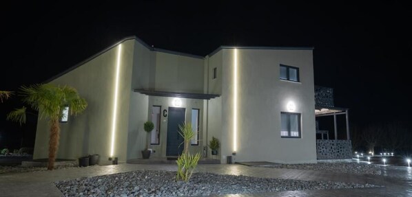 A modern two-story house with a well-lit entrance, a small garden area with rocks and plants, and a paved driveway. - The Rooms (Almopia)