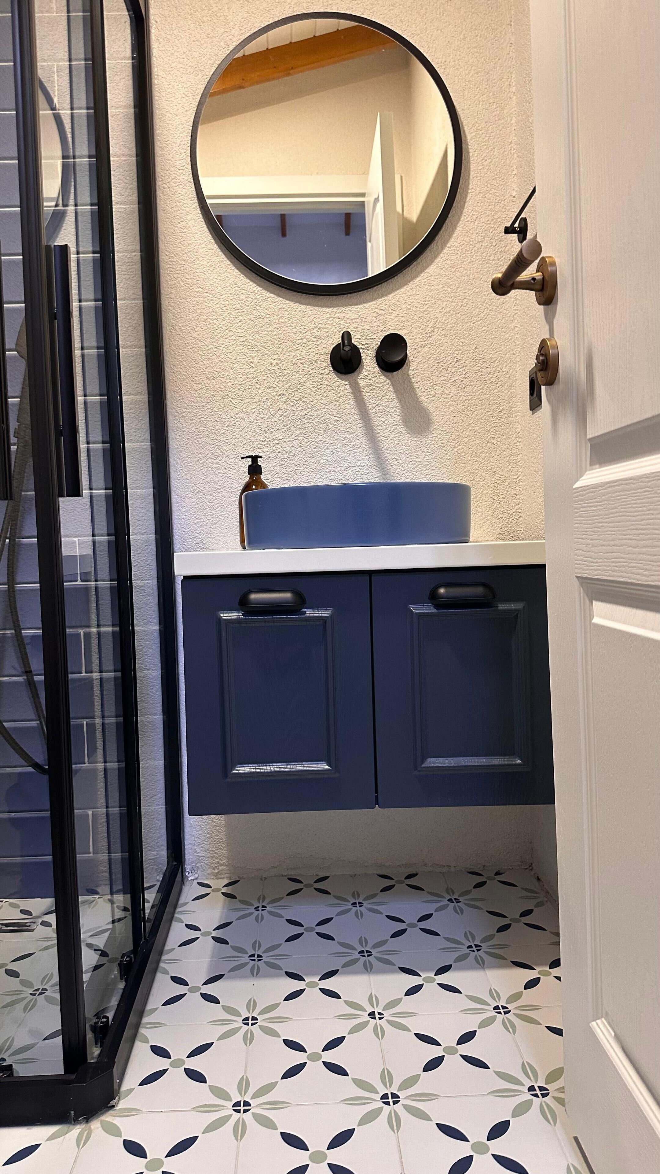 Baño con cabina de ducha de vidrio, espejo redondo, lavamanos con encimera azul y armarios azul oscuro.