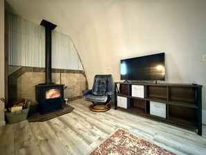 Una sala de estar con estufa de leña, un sillón de cuero, televisor sobre un mueble bajo y una estantería.