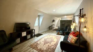 Un salon de style industriel avec un plafond haut, une grande fenêtre, un téléviseur à écran plat, un canapé foncé, un tapis à motifs et un luminaire moderne.