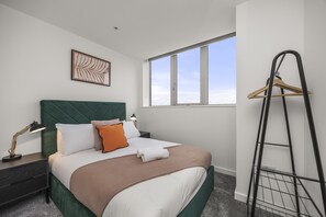 A neatly made bed with a green headboard, white and orange pillows, and a brown blanket. There is a bedside table with a lamp and a black metal rack against the wall. - Ocean View E By My Getaways (St Leonards-On-Sea)