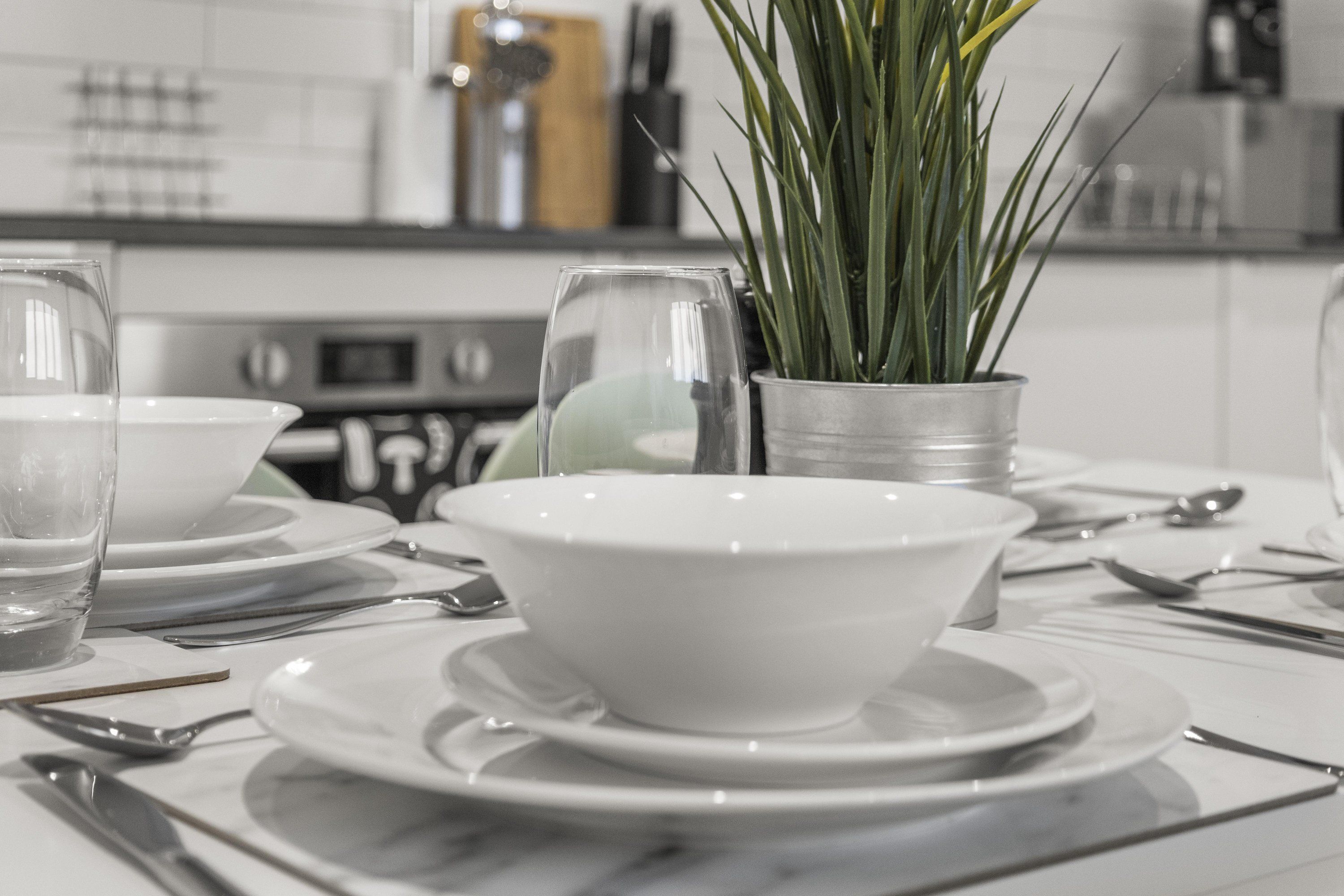 Una cocina con electrodomésticos blancos, una planta en maceta y una mesa de comedor lista con platos, tazones y cubiertos.