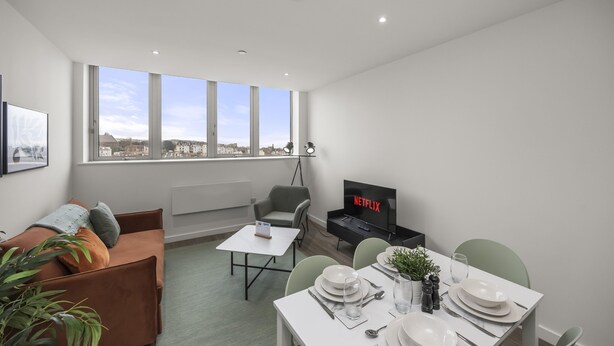Een moderne woonkamer met een bank, een eettafel gedekt voor twee personen, een televisie waarop 'NETFLIX' wordt weergegeven, en grote ramen met uitzicht op de stad.
