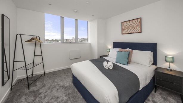 Ein modernes Hotelzimmer mit einem großen Bett, einem Schuhregal, einem Fenster mit Blick auf die Stadt und einem Nachttisch mit Lampe.