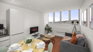 Ein modernes Wohnzimmer mit Essbereich, einem grauen Sofa, einem Fernseher und einem großen Fenster mit Blick auf die Stadt.