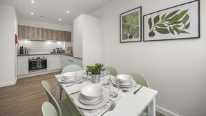 Un espace repas moderne avec une table blanche dressée pour quatre personnes, une cuisine en arrière-plan et deux reproductions botaniques encadrées accrochées au mur.