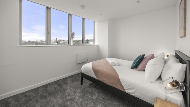 Une chambre moderne avec une grande fenêtre, un lit avec des oreillers, une table de chevet et une lampe fixée au mur.