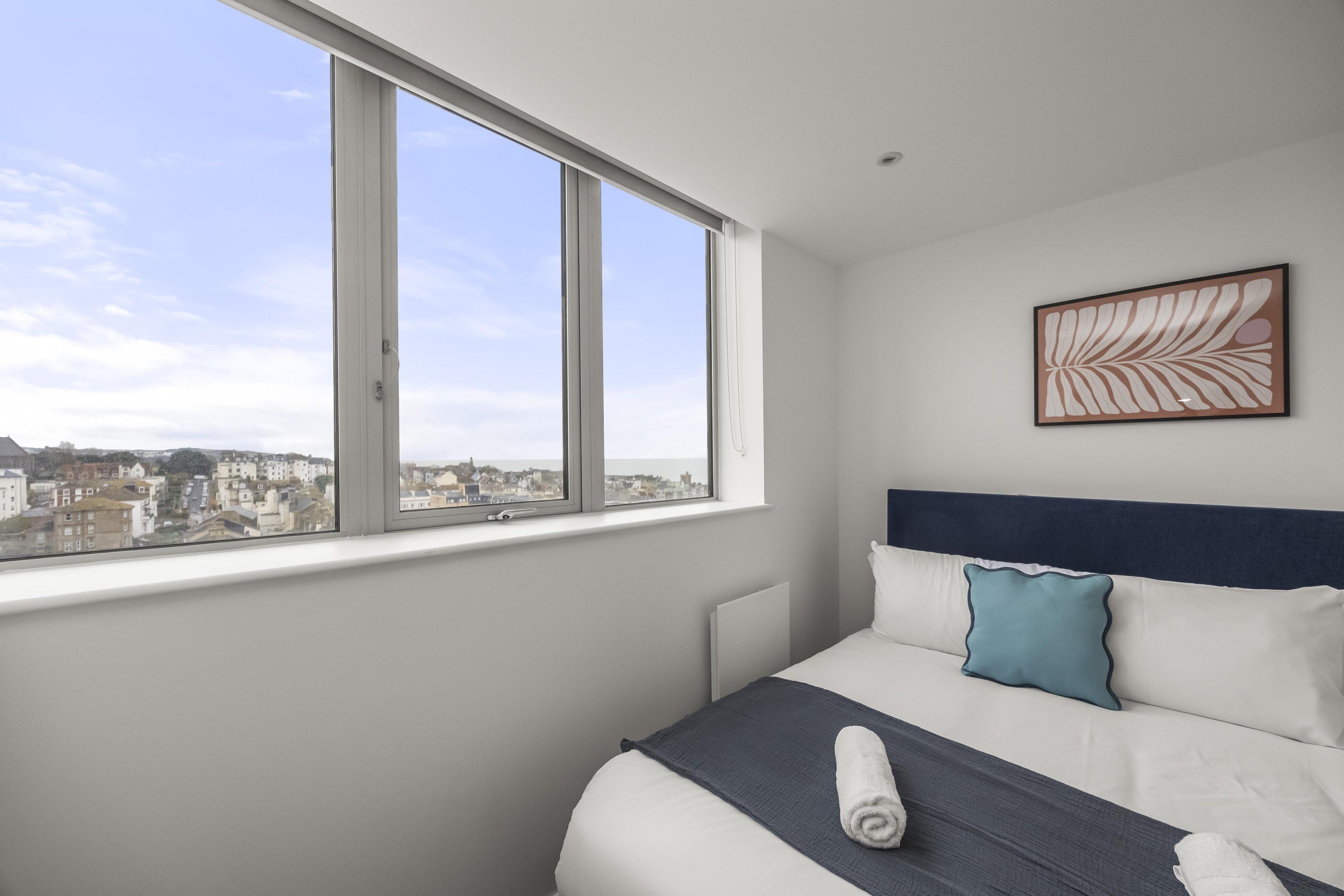 A hotel room with a large window, a bed with white bedding and a blue pillow, and a framed artwork on the wall.