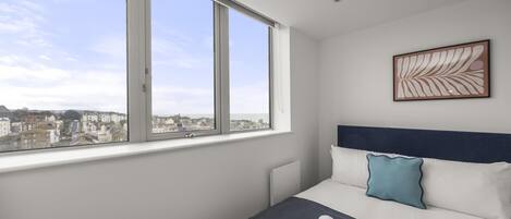 A hotel room with a large window, a bed with white bedding and a blue pillow, and a framed artwork on the wall.