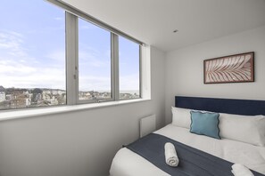 A hotel room with a large window, a bed with white bedding and a blue pillow, and a framed artwork on the wall.