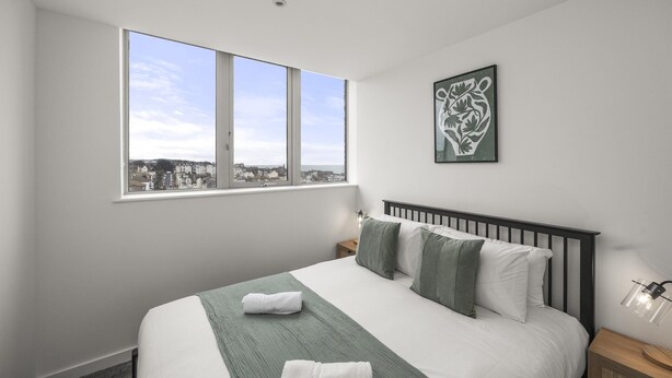 Una habitación de hotel con un ventanal grande, una cama con ropa de cama verde, una mesita de noche y una lámpara fijada a la pared.