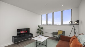 Una sala de estar moderna con un televisor de pantalla plana, una mesa de centro, un sofá, una silla, una planta y un ventanal con vistas a la ciudad.