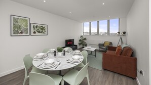Una sala de estar moderna con comedor, un sofá, un televisor y un amplio ventanal con vista a la ciudad.