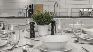 Una mesa de comedor bien puesta, con platos blancos, boles y vasos transparentes, acompañada de saleros de pimienta negra y una planta en maceta.