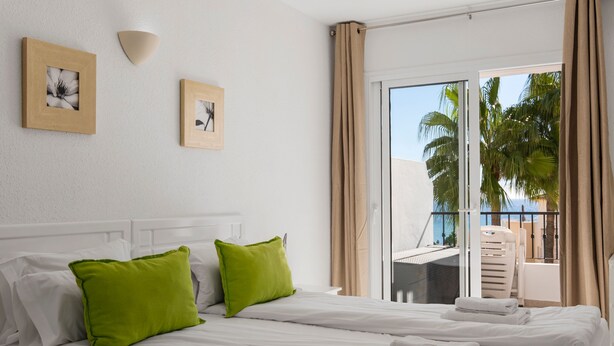 Une chambre d’hôtel avec un lit, deux oreillers, une lampe fixée au mur et une vue sur les palmiers, visible à travers une porte-fenêtre coulissante.