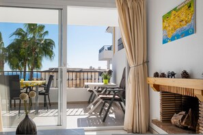 A balcony with a table and chairs, a potted plant, and a view of palm trees and a building across the water. - Sunny Flat With Pool in Riviera del Sol - Ref 96 (Mijas)