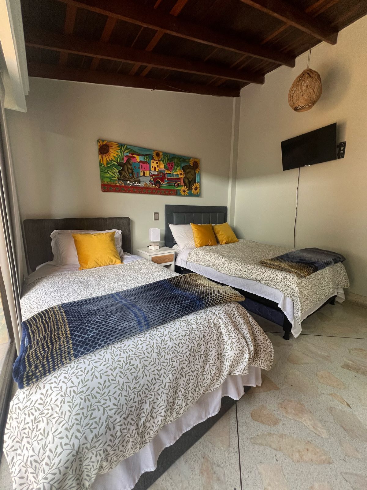 A room with two beds, each with patterned bedding and a headboard. There is a painting on the wall above the beds, a flat-screen TV mounted on the wall, and a woven basket hanging from the ceiling.