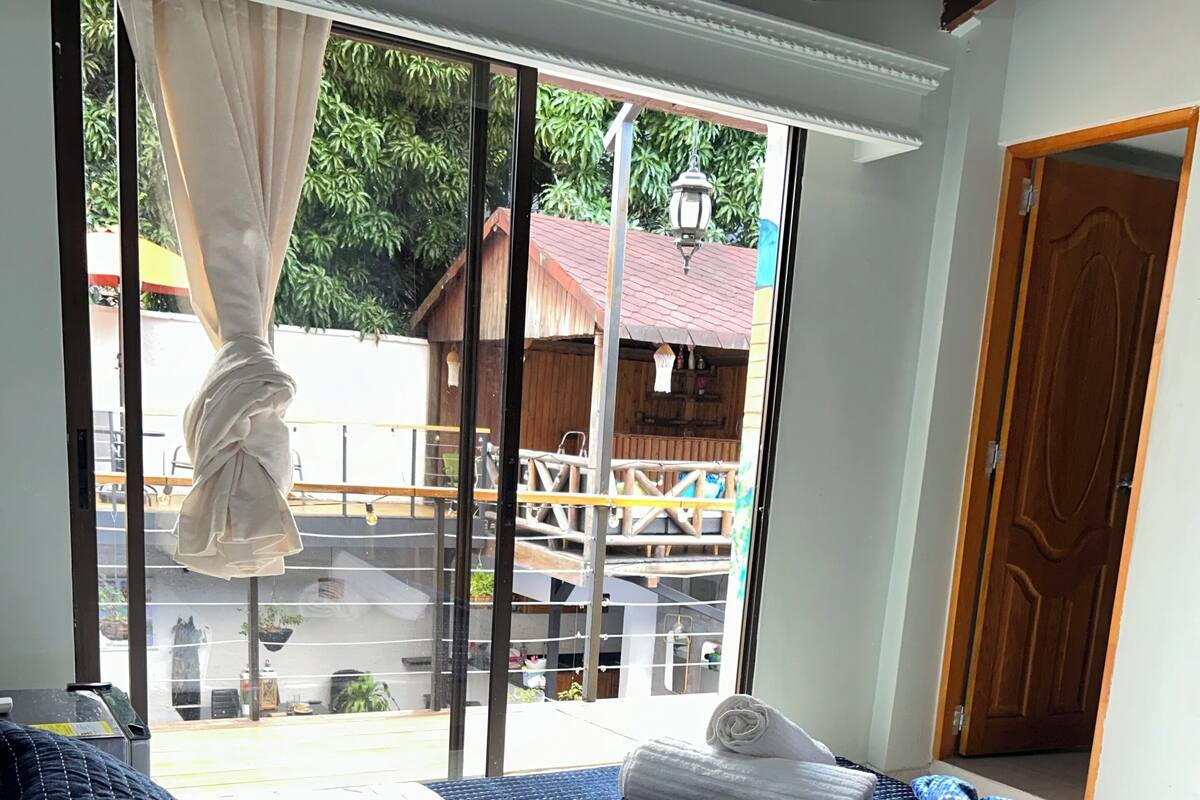 A room with a bed, a window with curtains, a wooden door, and a view of an outdoor area with a wooden structure.