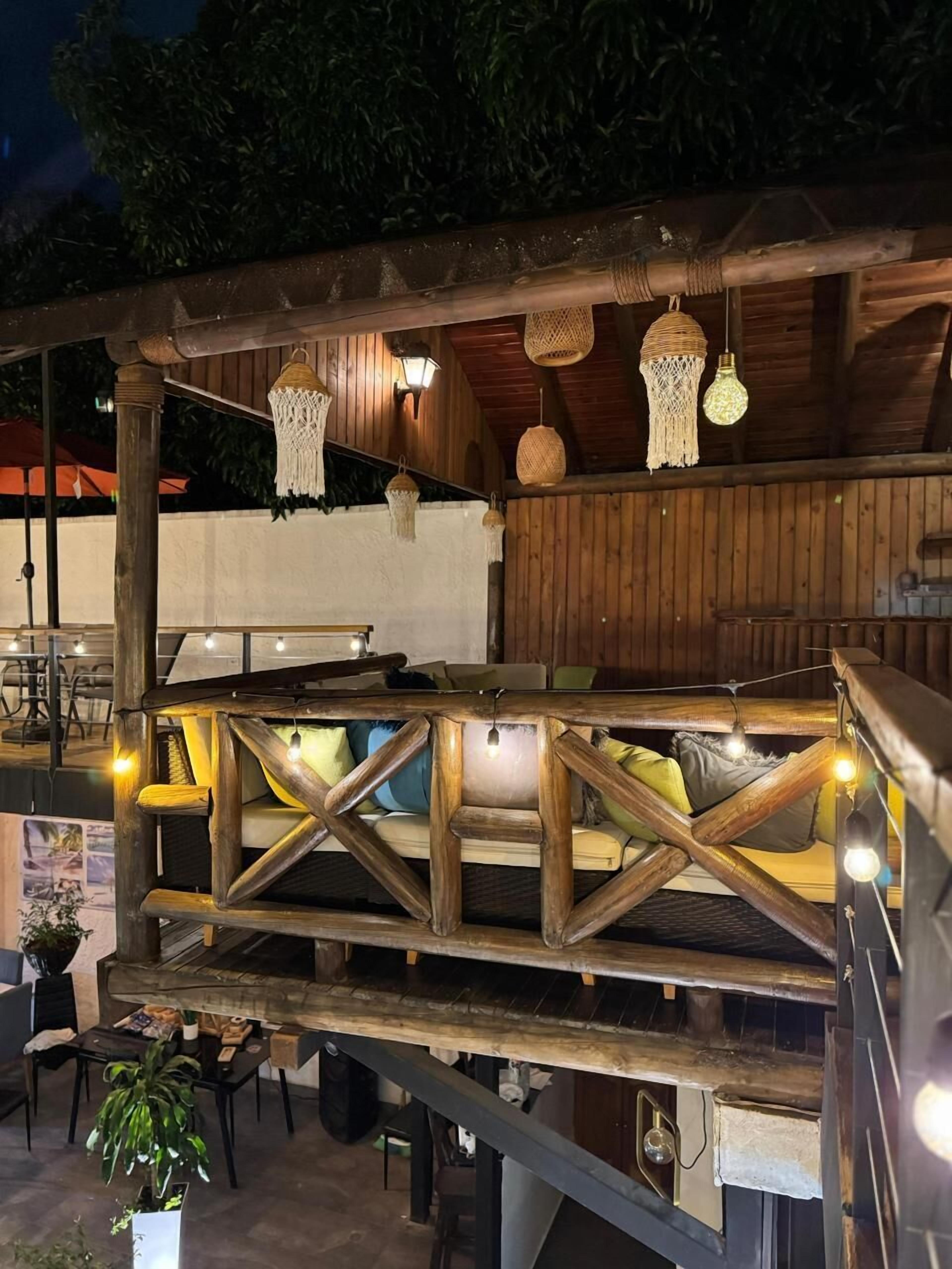 Una terraza de madera con barandilla, mesas, sillas y luces colgantes.